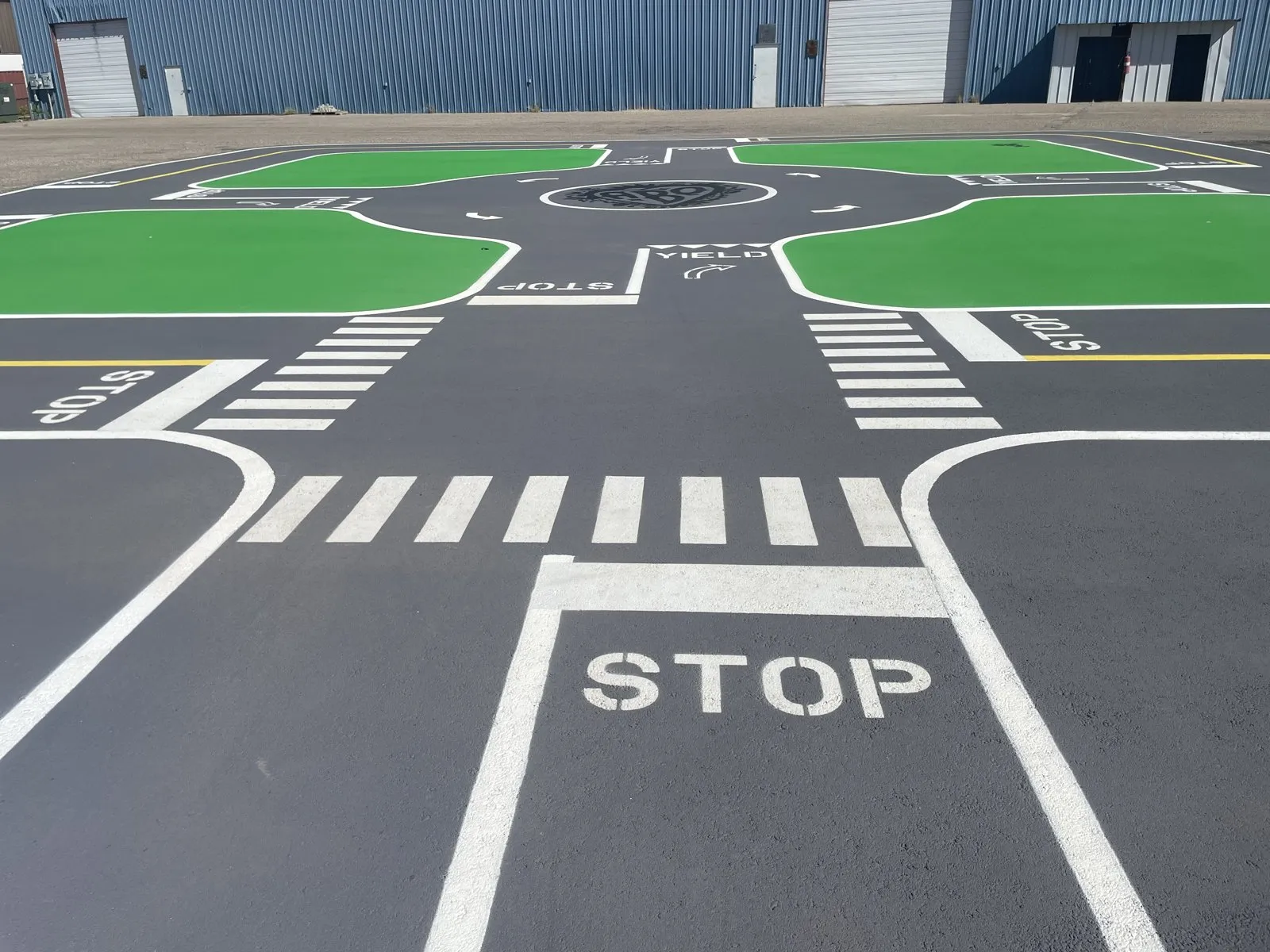 A kid-sized intersection painted on asphalt: a stop line and the word STOP, a crosswalk, and a small roundabout ahead between green corners