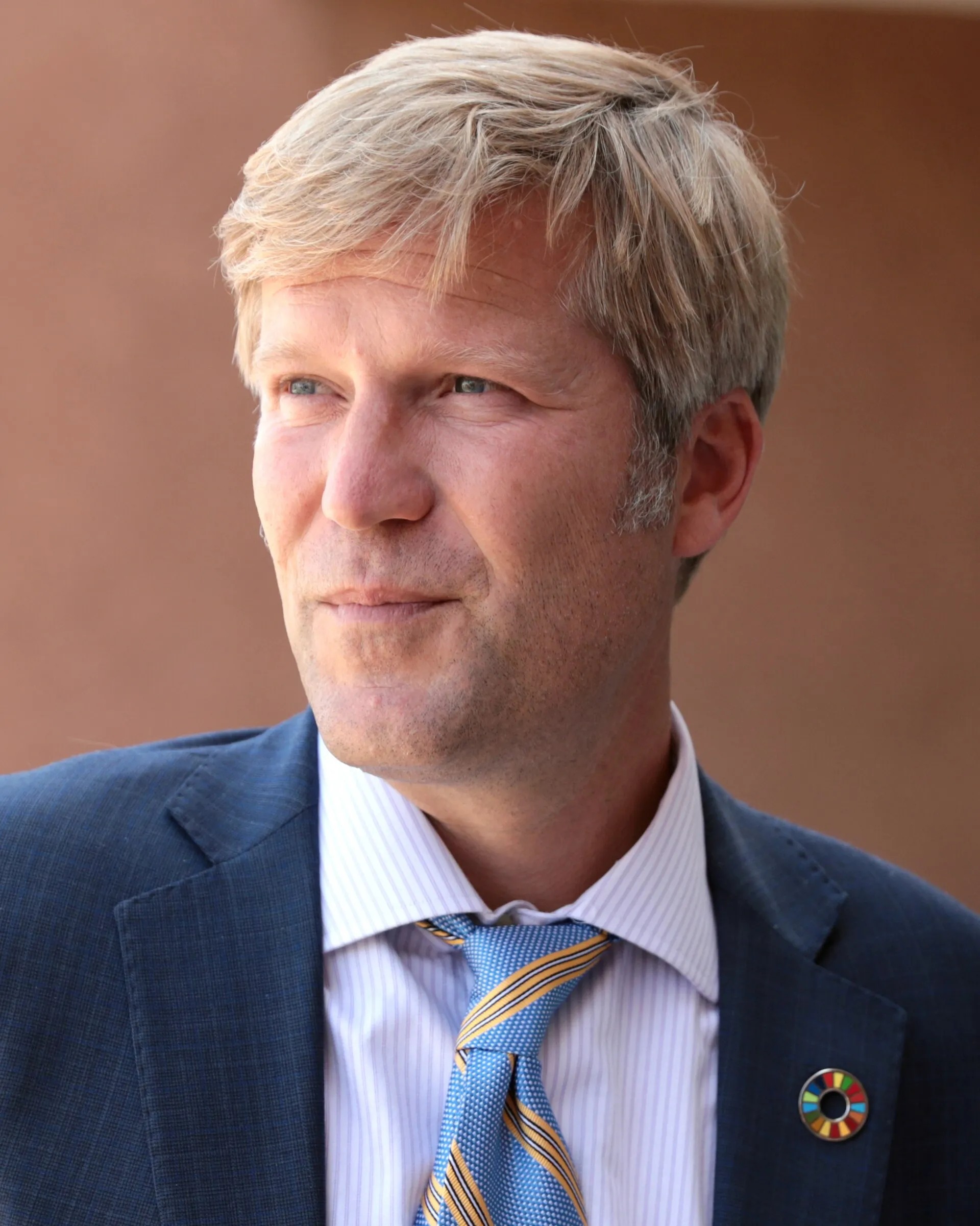 Tim Keller, a fair-haired man in a blue suit and striped tie, looking off to one side outdoors against an adobe wall